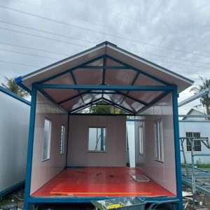 Fabrication stage of modular container classroom with pitched roof frame and red subfloor installation.
